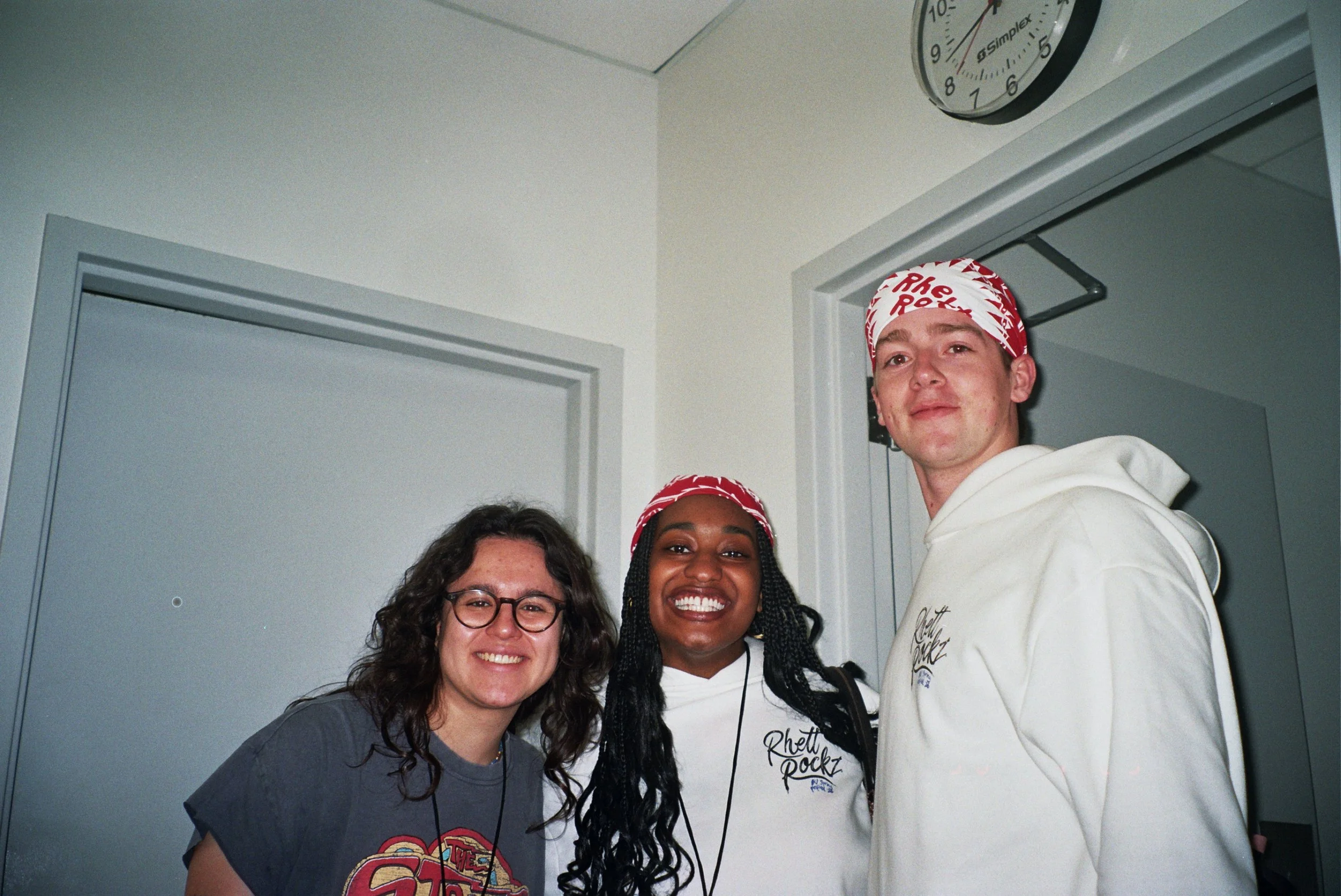 Group of four smiling women and a man standing closely together in an indoor setting, with two of them wearing red and white bandanas. A clock is visible on the wall in the background.