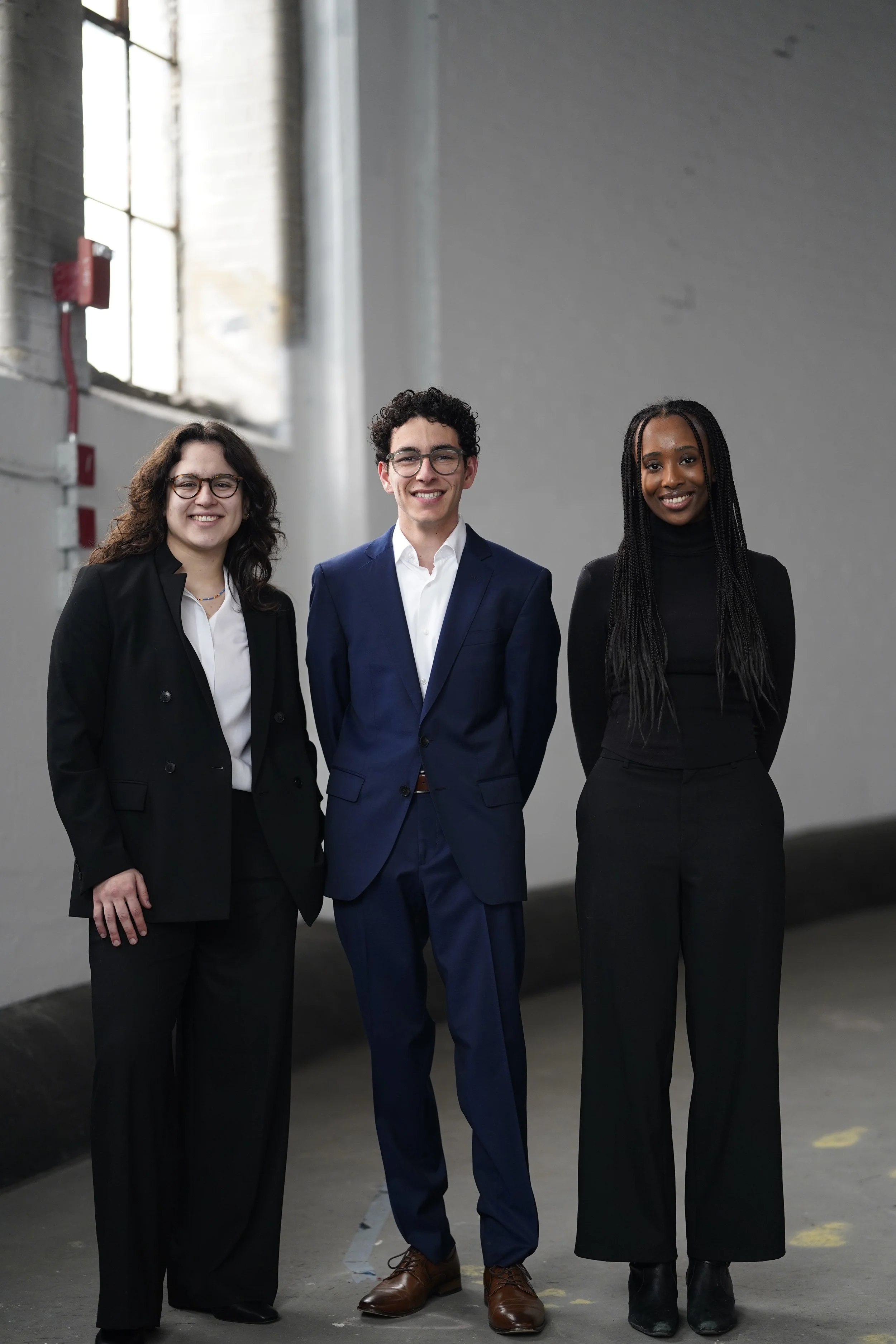 Three professionally dressed individuals standing together in an industrial or warehouse setting, smiling at the camera.