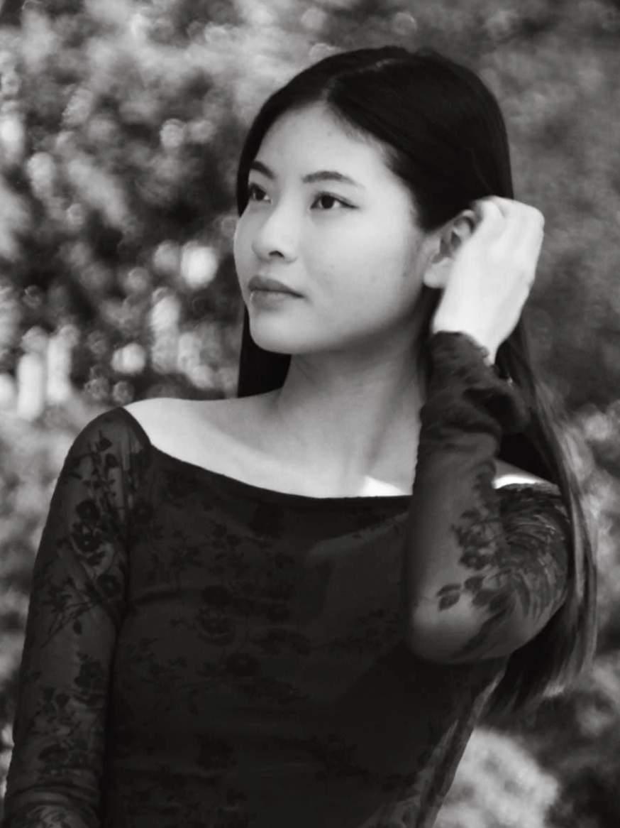A black-and-white portrait of a young woman with long dark hair, wearing a dress with lace sleeves, and touching her hair with her right hand, outdoors with blurred trees in the background.