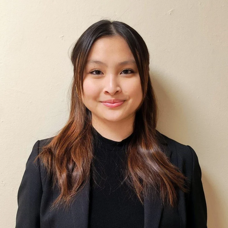 Young woman with long brown hair wearing a black blazer and top, standing against a plain beige wall, smiling gently at the camera.