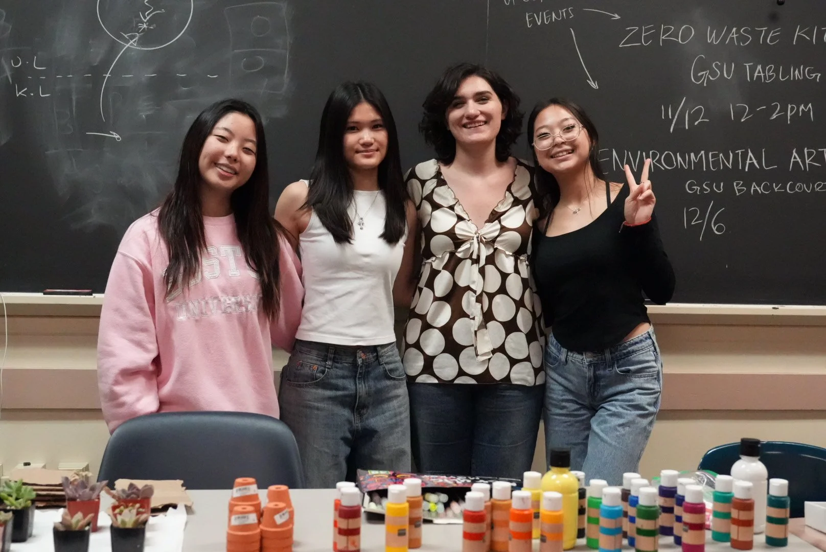 Four women standing together in a classroom with a blackboard behind them, smiling. The blackboard has handwritten notes about environmental art events. The table in front has various art supplies like paints, brushes, and craft materials.