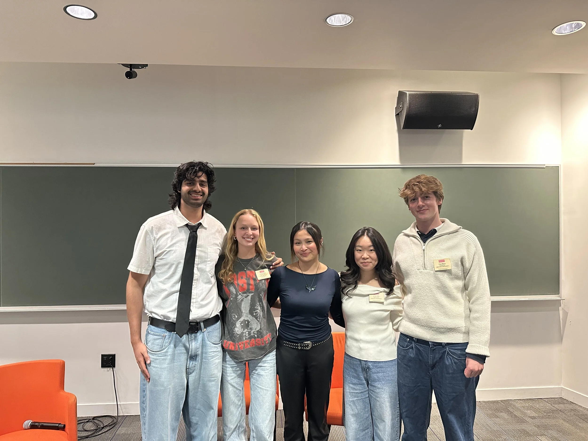 Group of six young adults standing together in a classroom or conference room, smiling and looking at the camera.