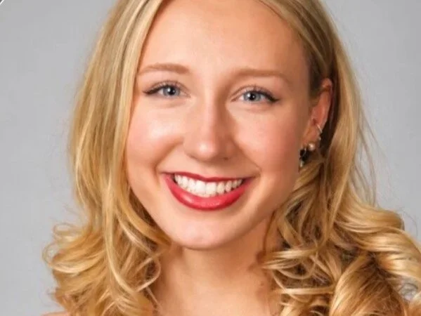 A young woman with blonde hair, red lipstick, and earrings smiling at the camera against a gray background.