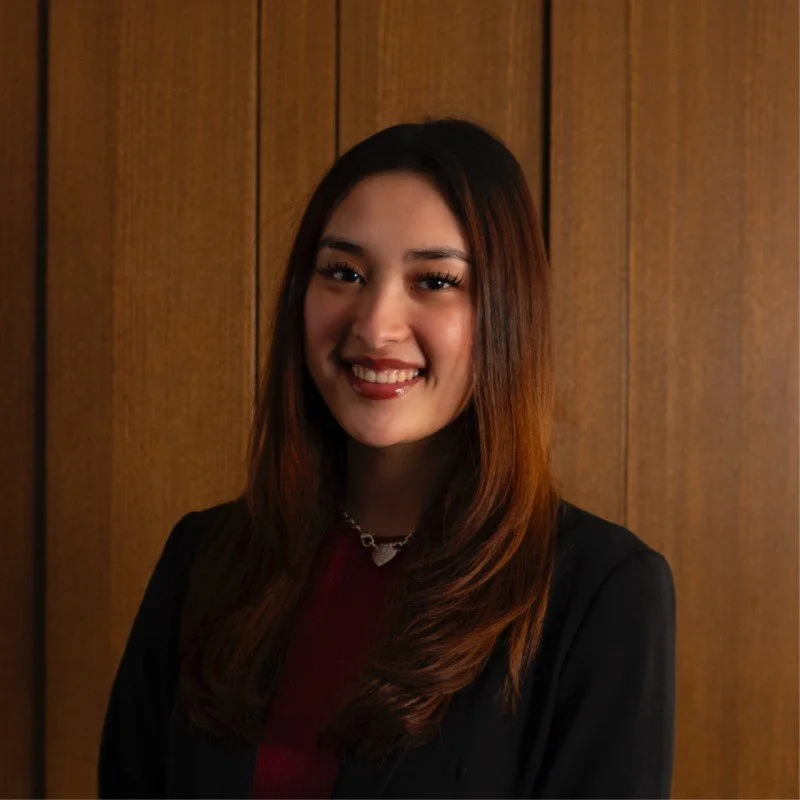A young woman with long brown hair, smiling, wearing a black blazer and a necklace, standing against a wooden-paneled background.