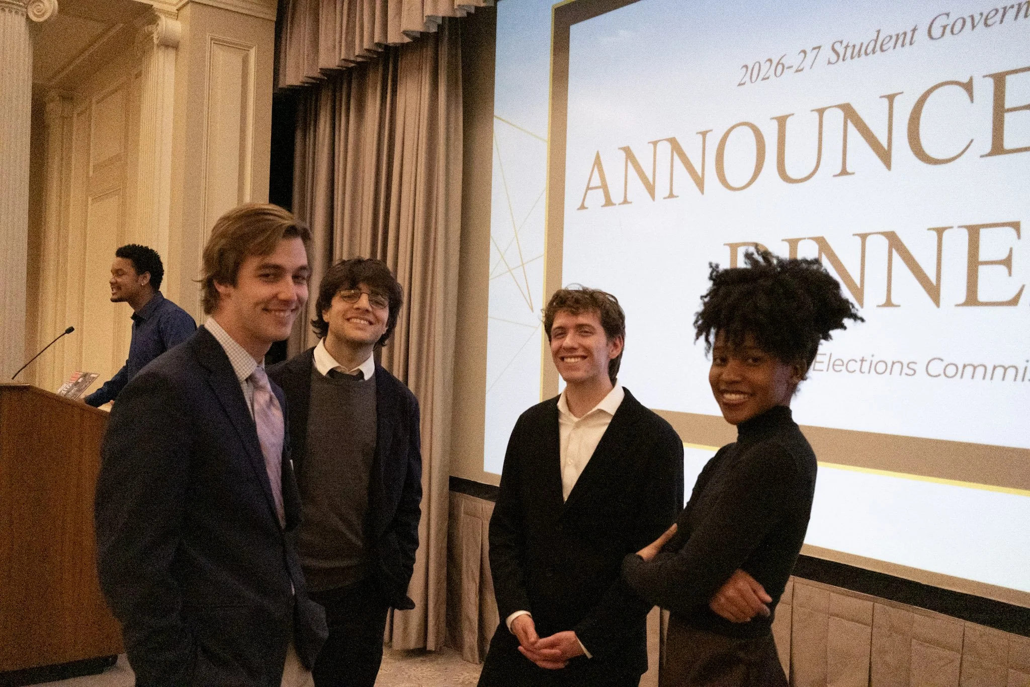 Group of five young adults in formal attire standing in front of a large presentation screen with the words '2026-27 Student Governance' and 'Announcement' displayed. They are smiling and posing for the photo at an indoor event.