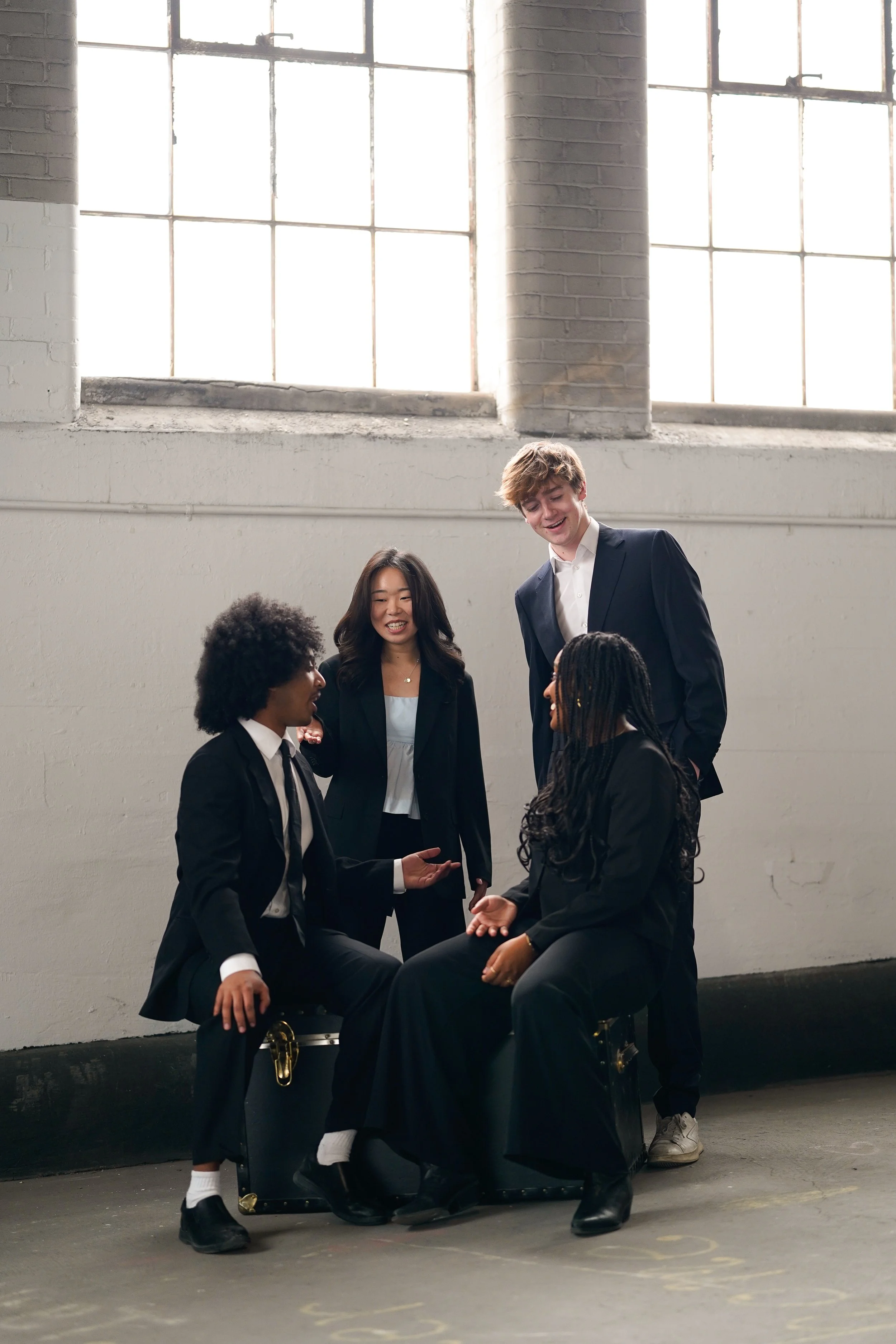 Six young adults, three men and three women, are gathered in a room with large windows and white painted brick walls. They are dressed in formal attire and are engaging in a conversation, with some standing and some sitting, around a seated woman.