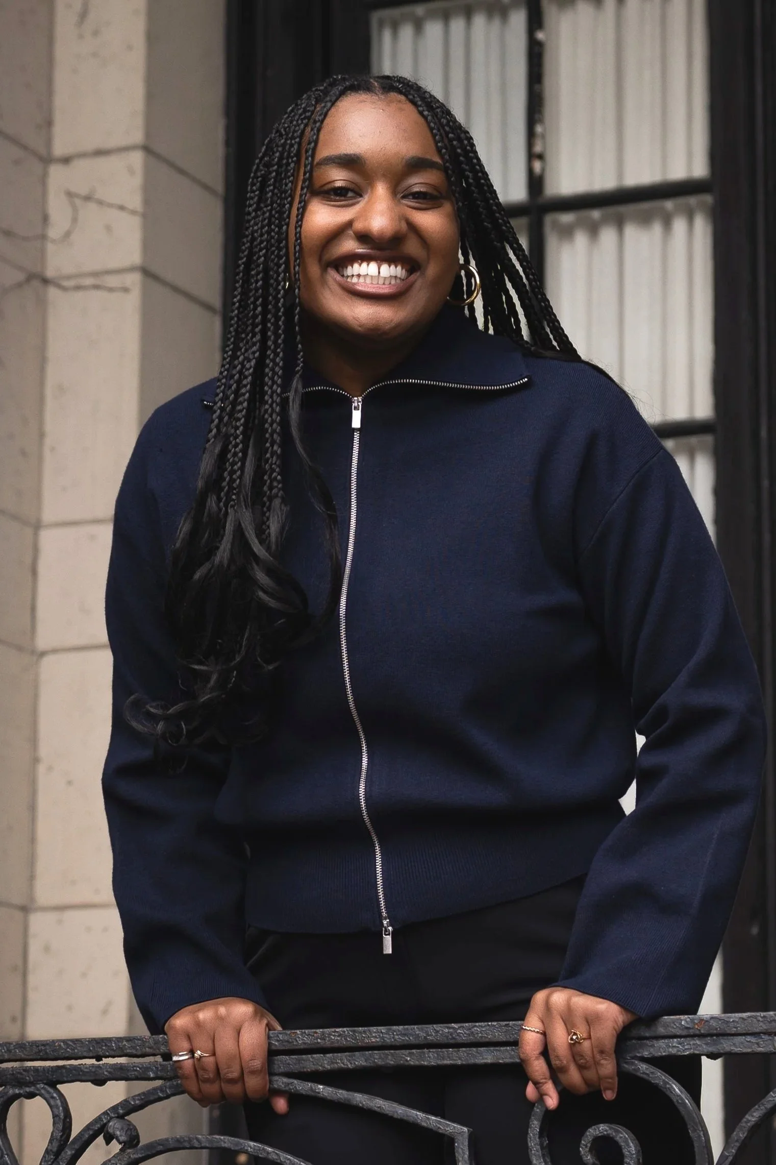 A smiling woman with long braided hair holding onto a decorative metal fence, standing in front of a building with stone and window frames.