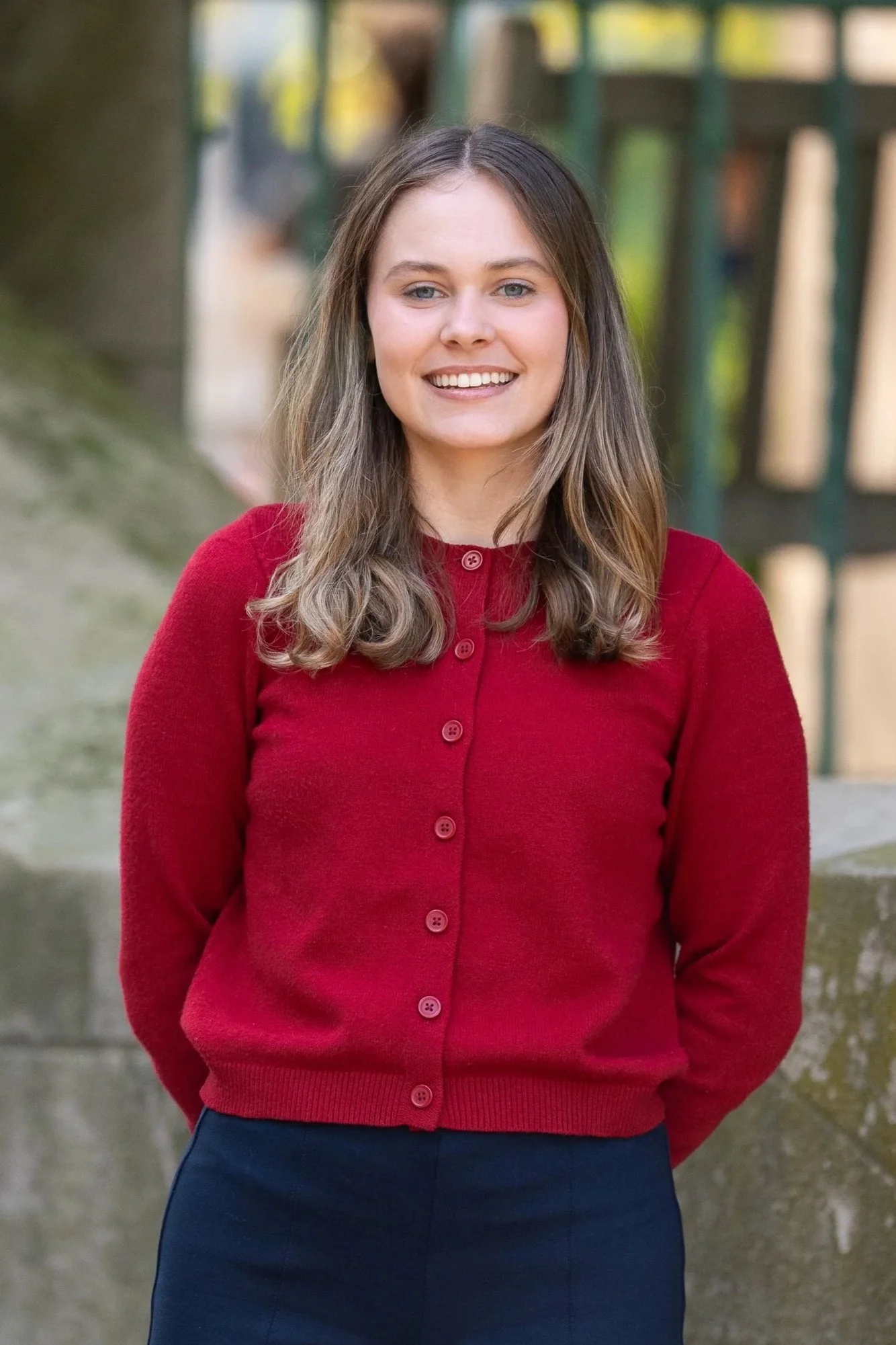 A woman with long, light brown hair and fair skin is smiling at the camera. She is wearing a red cardigan buttoned up, and dark pants. The background is outdoors with blurred greenery and a fence.