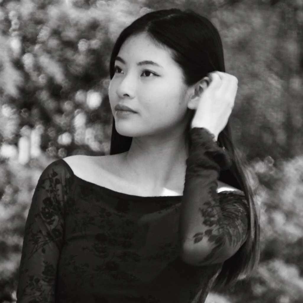 Black and white portrait of a woman with long dark hair, wearing a lace top with long gloves, standing outdoors with blurred foliage in the background.