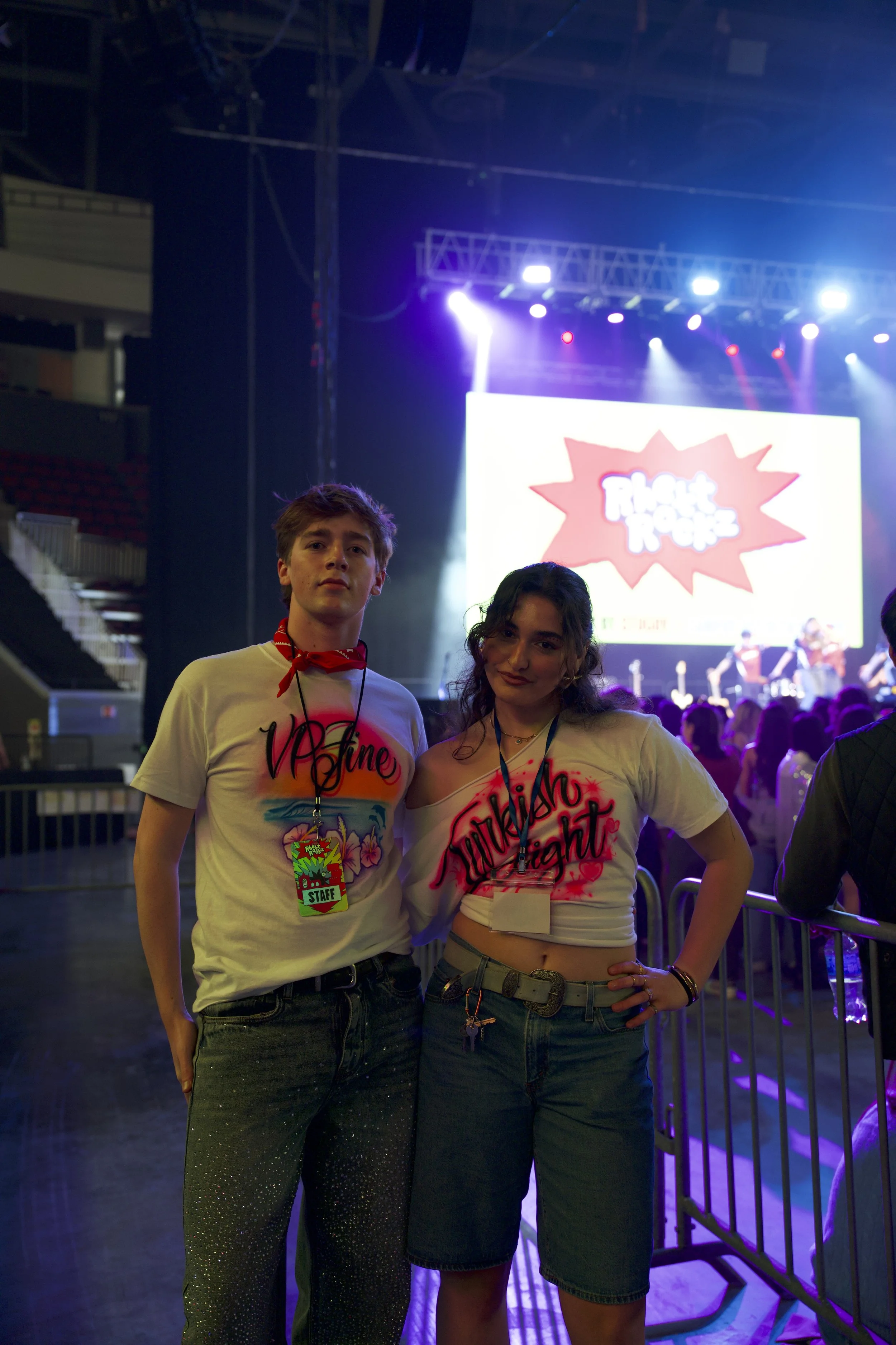 Two young women standing together at a concert, with bright stage lights and a large screen displaying a logo behind them. They are wearing casual clothes with event lanyards.
