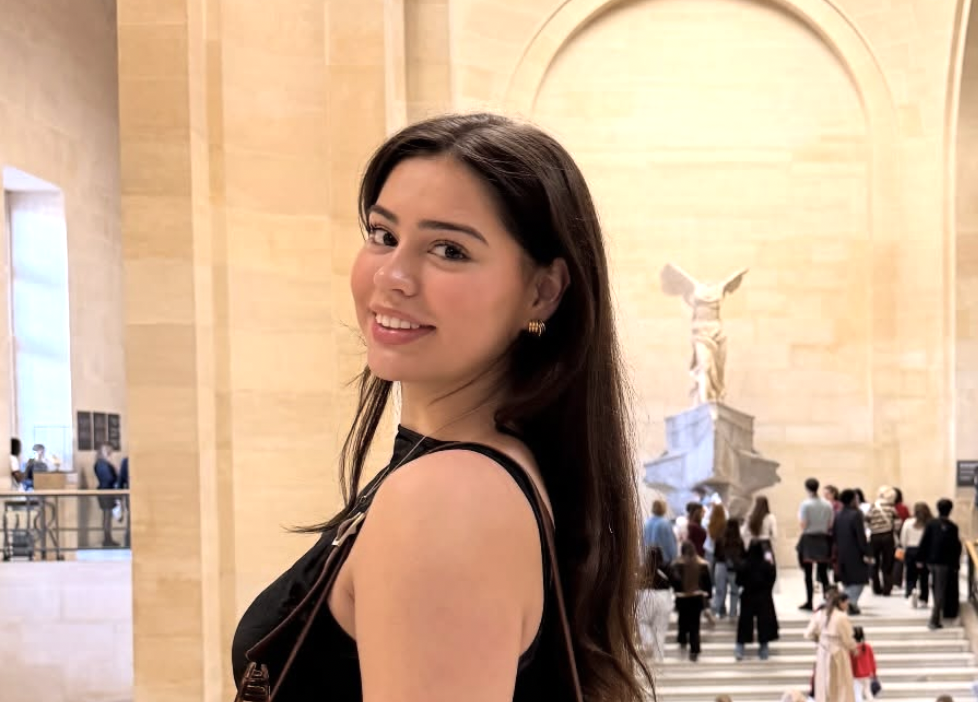 A young woman with long dark hair smiling at the camera inside an art museum, with a large statue and visitors in the background.
