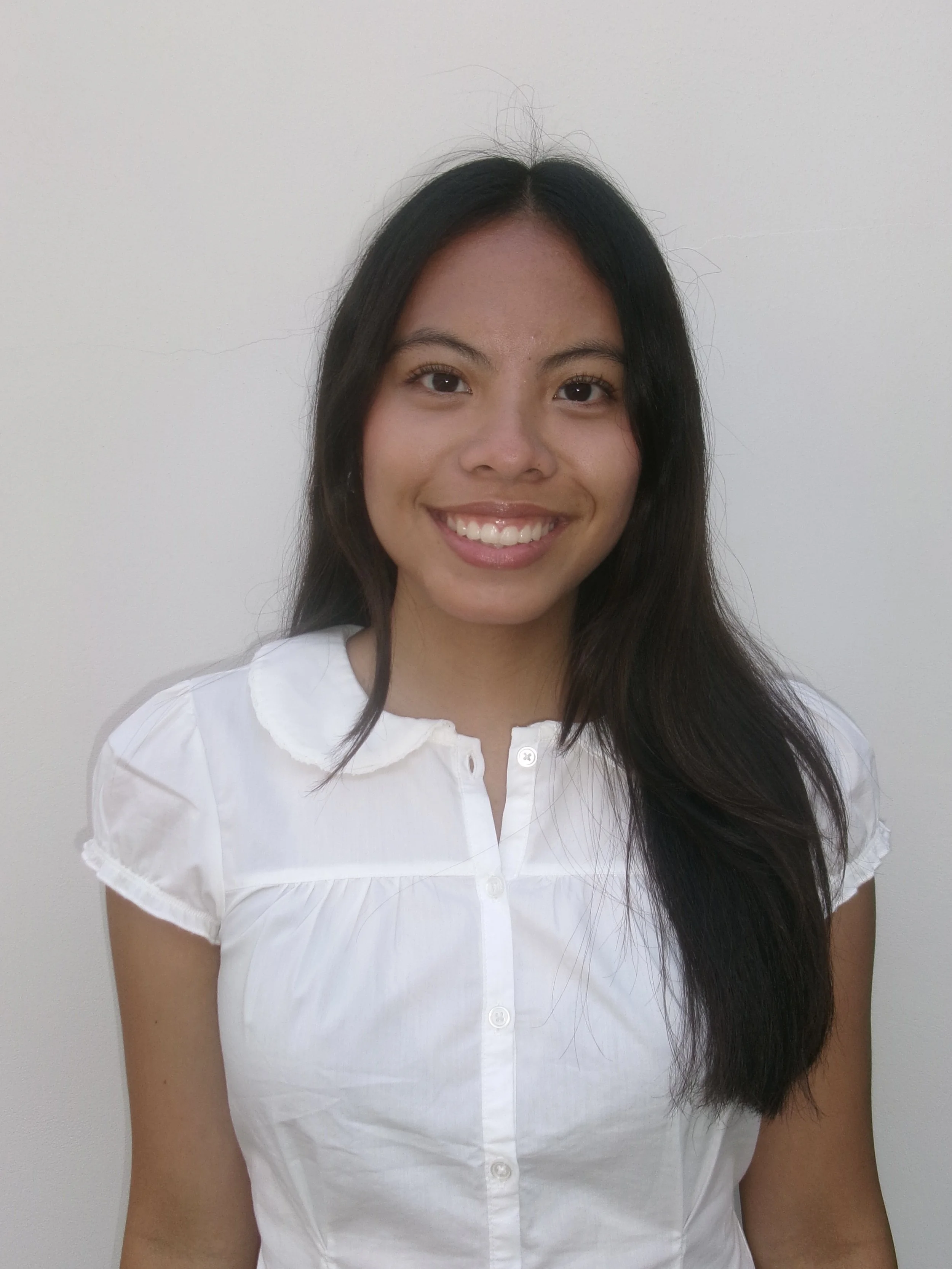 Close-up of a woman with long, dark hair wearing a white top, smiling against a plain light-colored background.