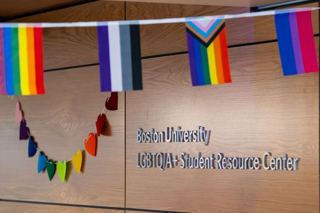 Sign on a wooden wall reading "Boston University LGBTQIA+ Student Resource Center" with rainbow flags and heart-shaped paper decorations hanging above.