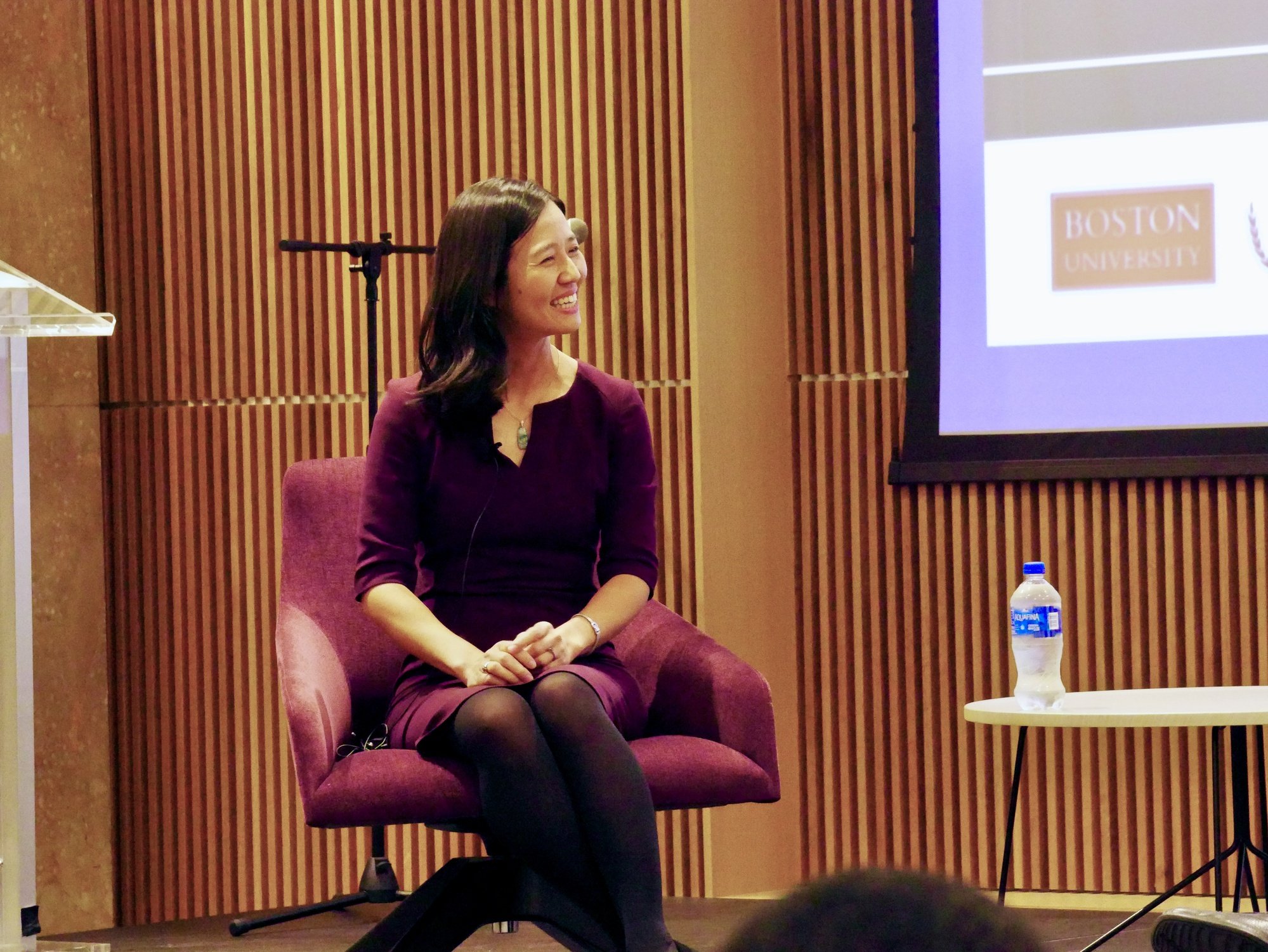 A woman sitting in a purple armchair during an event at Boston University, smiling and looking to her left. There is a water bottle on a small table next to her and a large screen in the background.