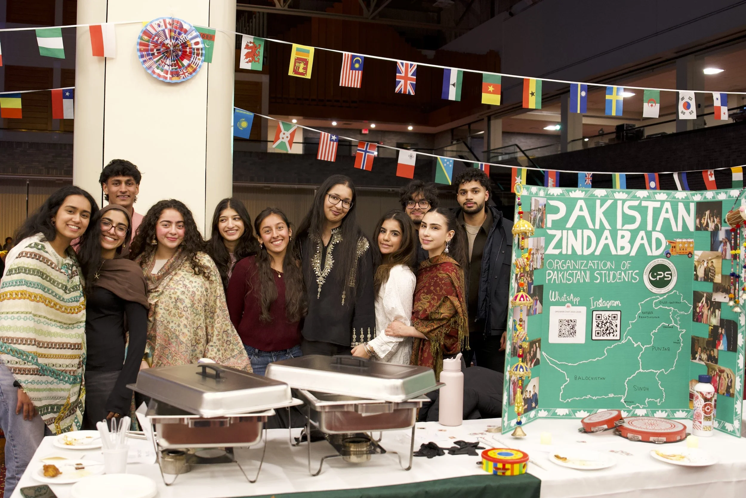 Group of young people standing in front of a booth with a Pakistan map, flags, and decorations, at an international cultural event.