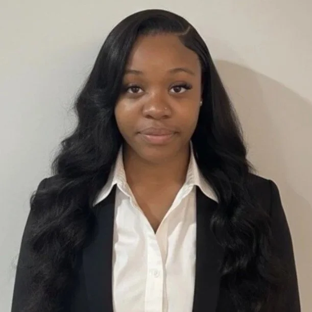 Young woman with long black wavy hair wearing a white collared shirt and black blazer standing against a plain light-colored wall.