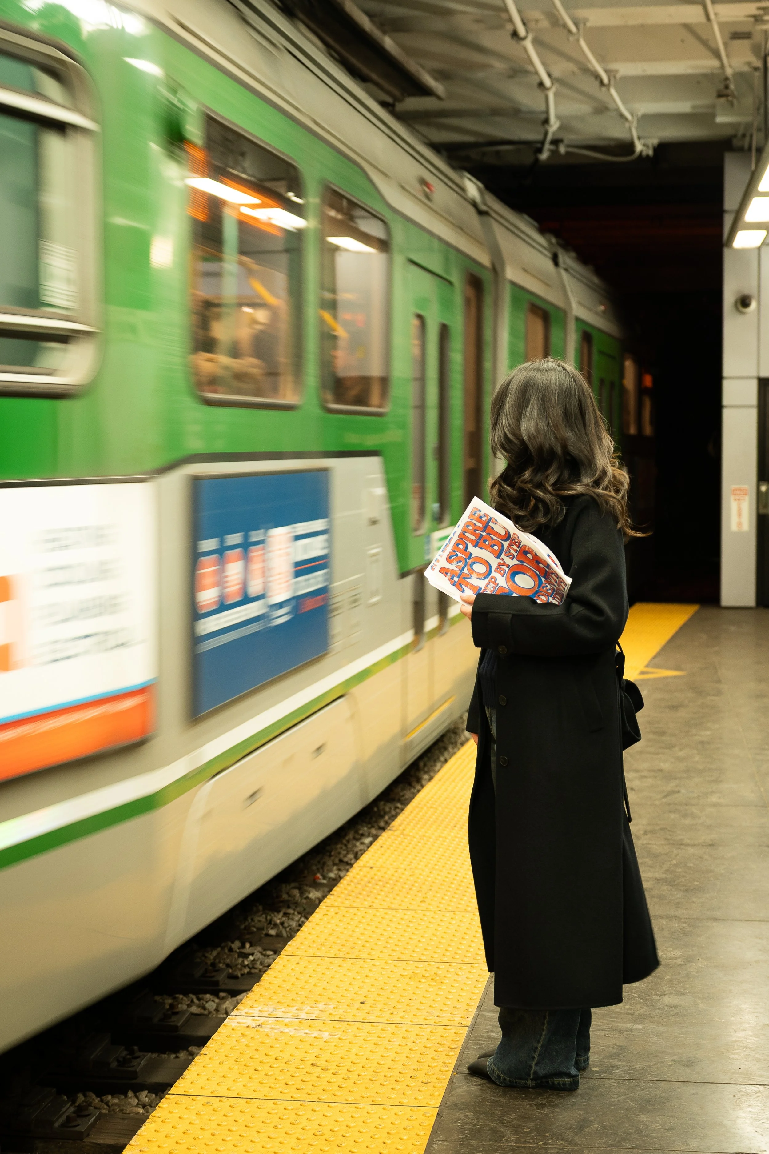 A woman with dark, wavy hair wearing a long black coat and jeans is waiting on a yellow platform at a subway station. She is holding a colorful magazine or brochure in her hand. A green train is arriving or departing behind her, and the station has black walls with some white paneling and a security camera visible in the corner.
