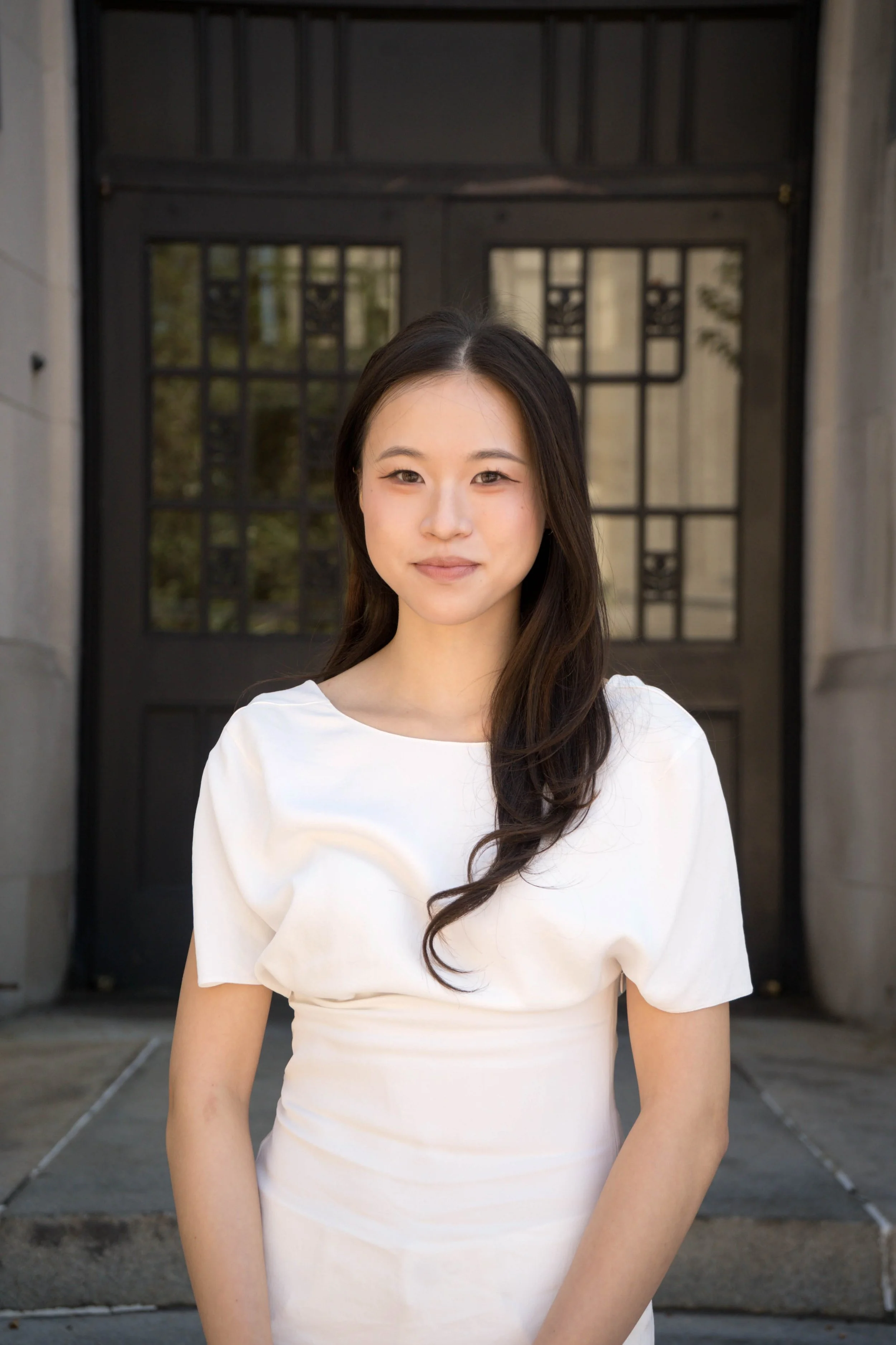 Portrait of a young woman with medium-length black hair, wearing a black blazer, looking directly at the camera with a slight smile, against a plain light background.