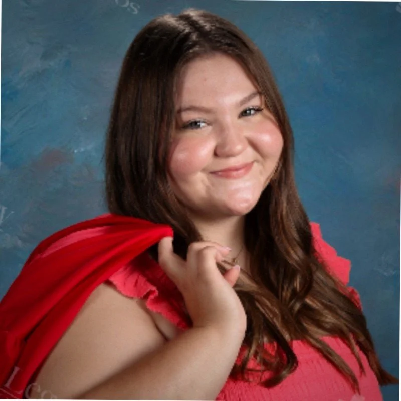 A young woman with shoulder-length brown hair smiling while holding a red cape over her shoulder, against a blue background.