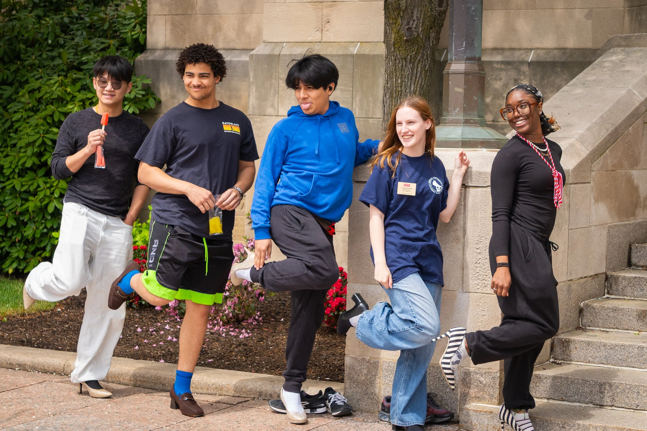 Five young people, four men and one woman, are standing outside near a stone building and smiling. Their poses suggest they are friends or participating in an event, with one person playfully sticking out his tongue and another holding a popsicle.