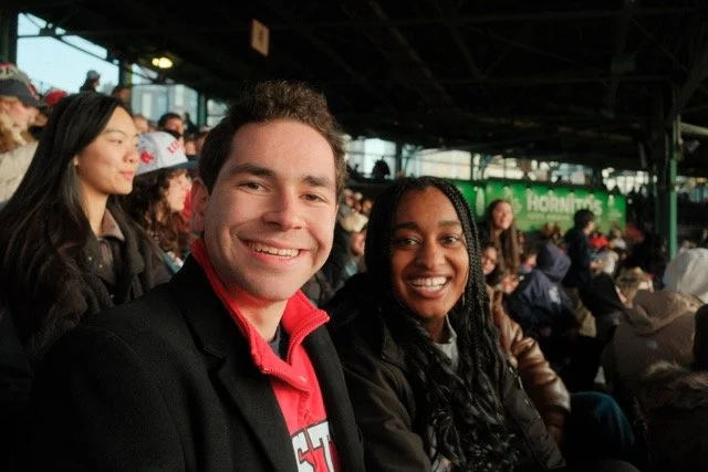 Two smiling people sitting in a crowded outdoor stadium or arena with other spectators in the background.