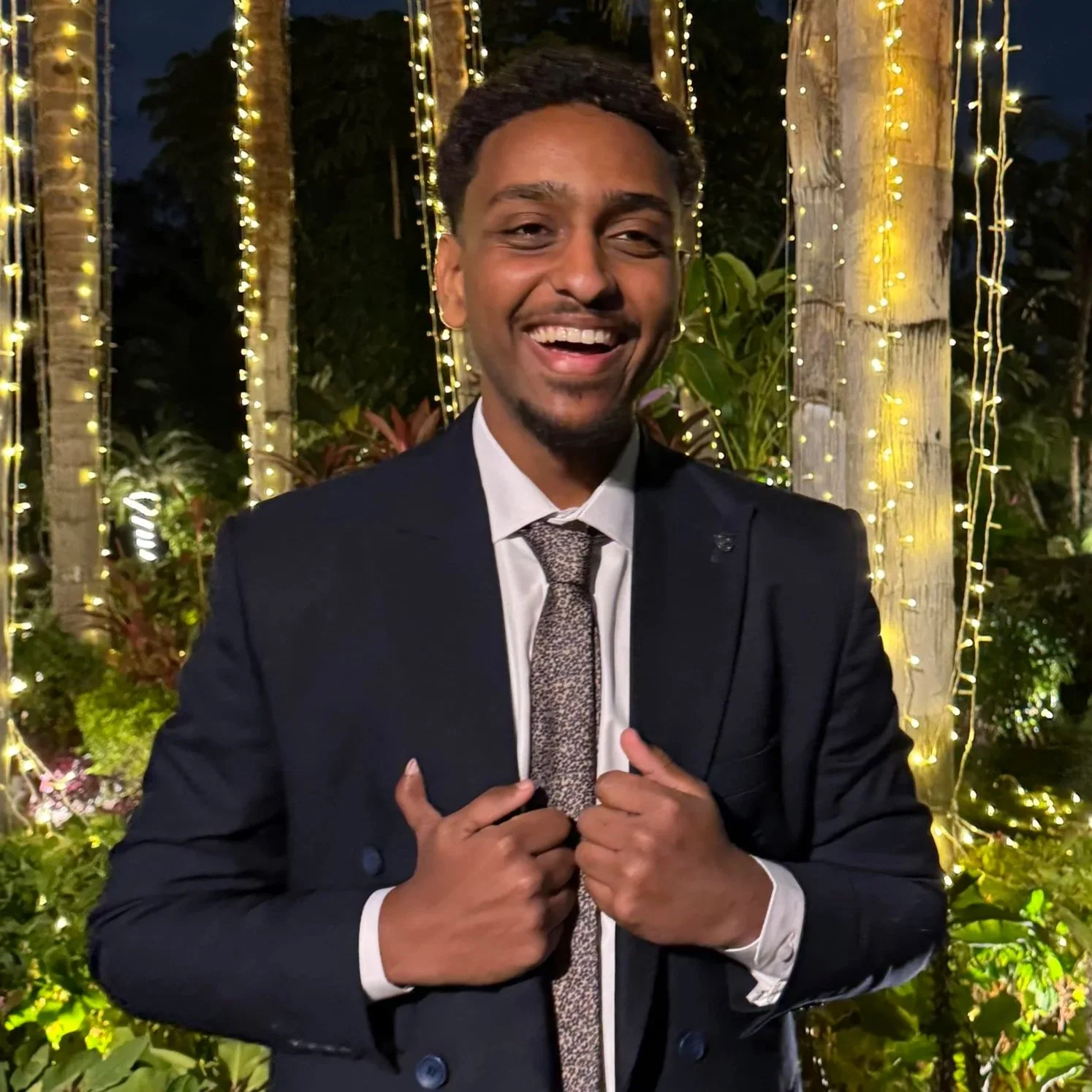 A smiling man in a suit and tie, standing outdoors at night, with string lights and trees in the background.