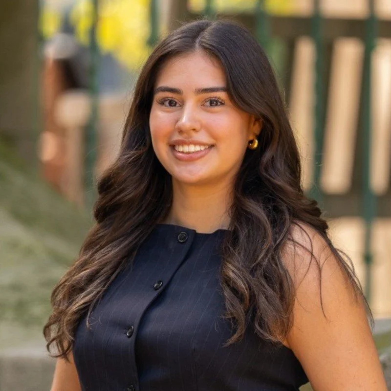 A young woman with long, wavy dark hair smiling outdoors, wearing a sleeveless navy blue top and gold earrings.