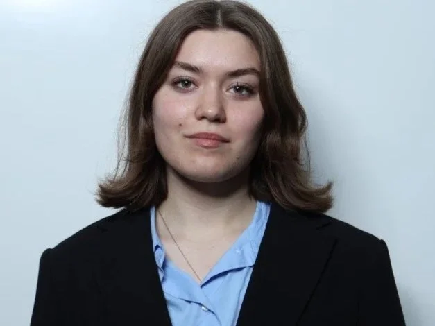 A young woman with shoulder-length brown hair wearing a black blazer over a blue collared shirt standing against a plain background.