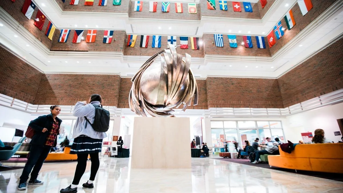 Interior of a modern building with a large, abstract metal sculpture in the center. Two women are in the foreground, one with a backpack. Several people are seated and walking in the background, with multiple flags hanging from the upper floors.