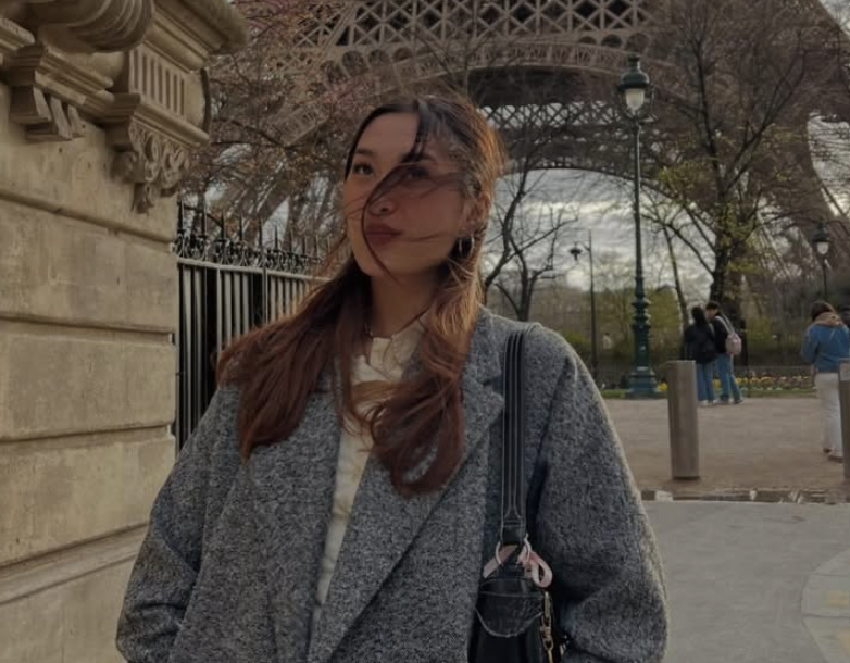 Young woman standing outdoors near a stone wall and fence, with trees, street lamps, and a structure resembling the Eiffel Tower in the background.