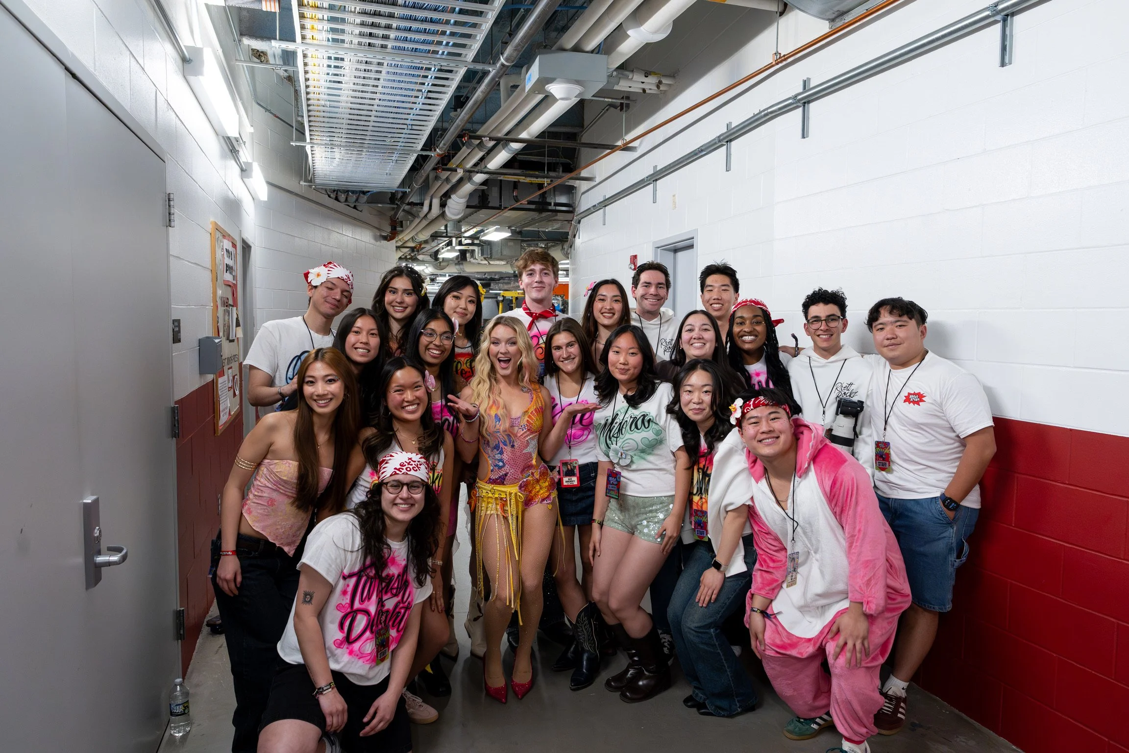 A diverse group of young adults dressed in colorful, tropical-themed clothing and accessories, including leis, floral headbands, and bright outfits, posing together in a hallway with white and red walls, ceiling piping, and a metal vent overhead, smi