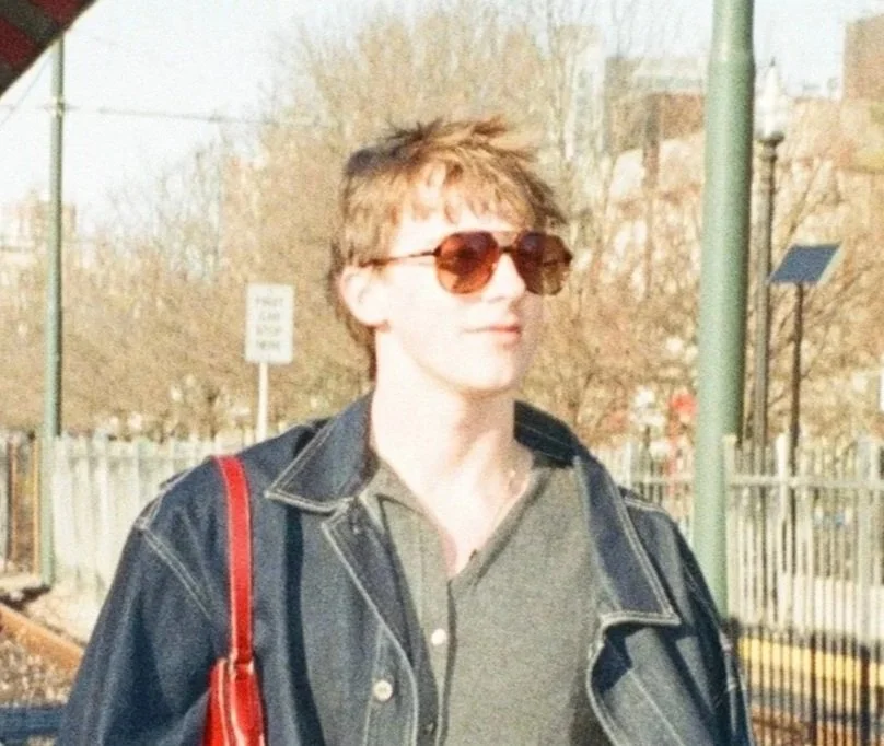 A woman with short, spiky blonde hair wearing brown sunglasses, a jean jacket, and a gray shirt, carrying a red bag, standing outdoors with trees and a fence in the background.