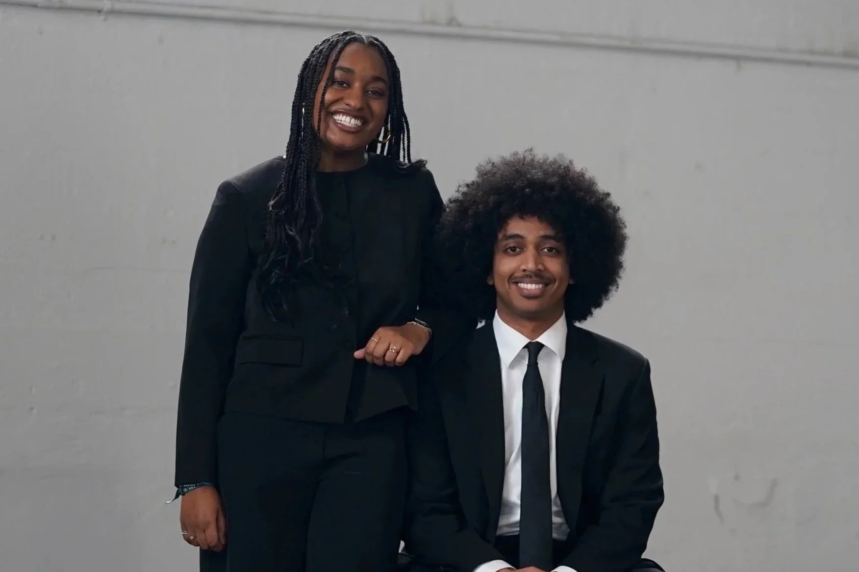 A woman standing next to a man seated, both dressed in black suits with white shirts and black ties, smiling against a plain gray background.