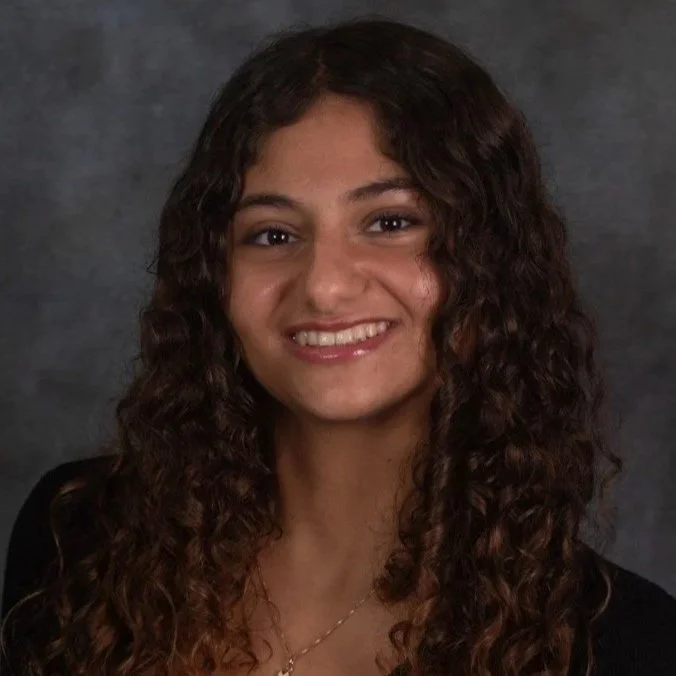 A woman with long, curly brown hair and a bright smile, wearing a black top and a gold necklace, against a gray backdrop.