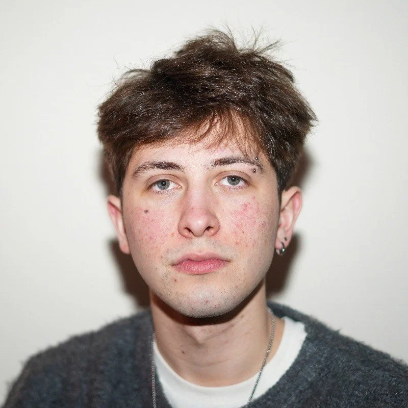 A young man with light skin, brown hair, and light-colored eyes looks directly at the camera. He has some acne on his face, wears a white shirt with a dark sweater over it, and a small earring in his left ear.