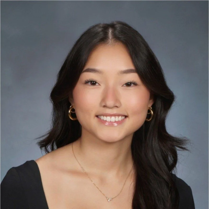 A young woman with long dark hair smiling at the camera, wearing a black top, gold hoop earrings, and a delicate gold necklace against a gray background.