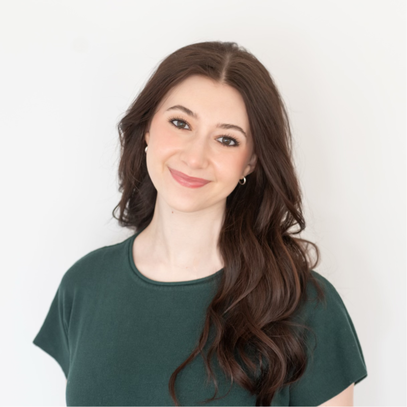 A woman with long, wavy brown hair and a gentle smile wearing a dark green shirt, standing against a plain white background.