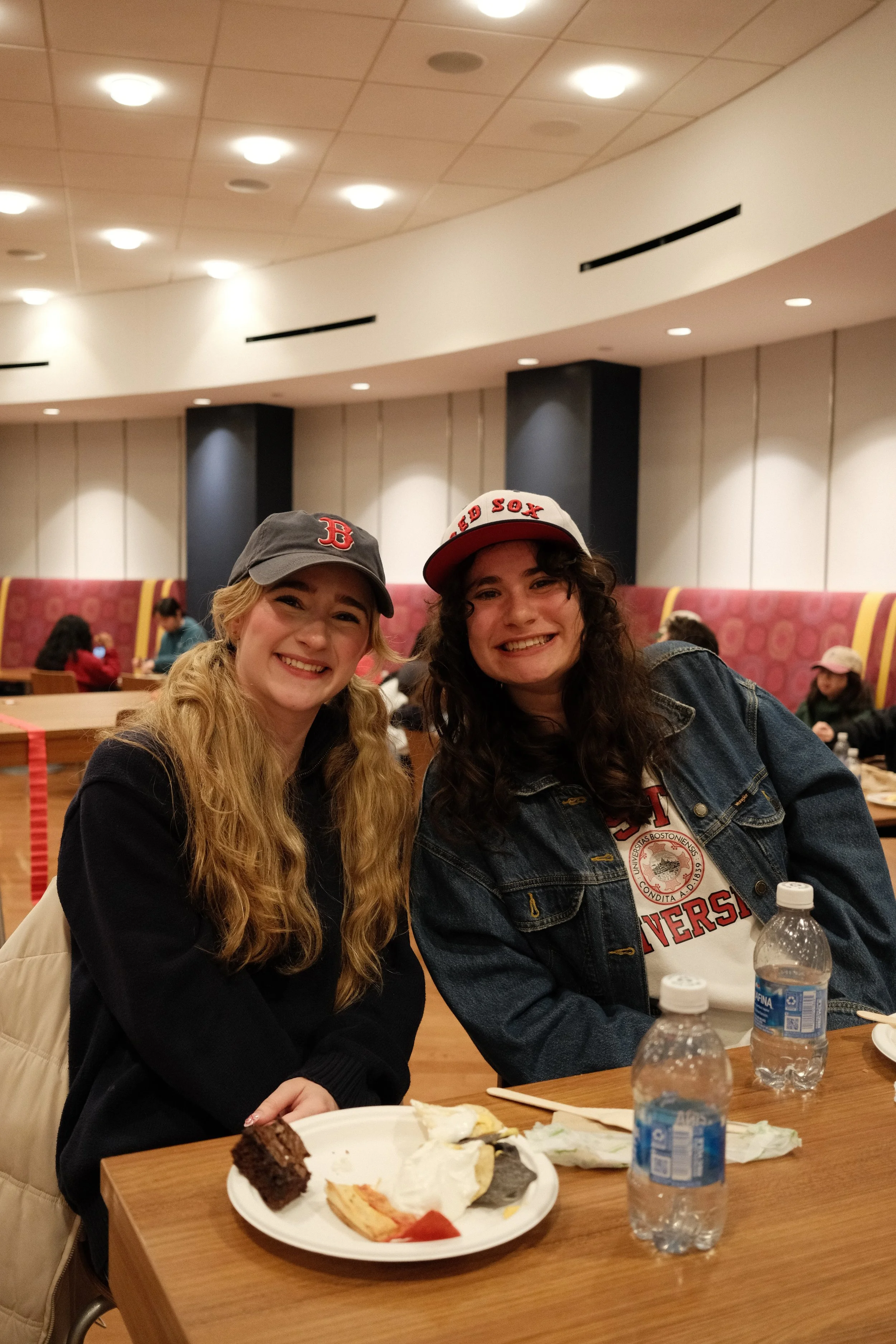 Two young women sitting at a table in a cafe or restaurant, smiling at the camera. They are wearing hats supporting the Boston Red Sox and Harvard University. The table has some leftover cake and two bottles of water, with other people visible in the background.