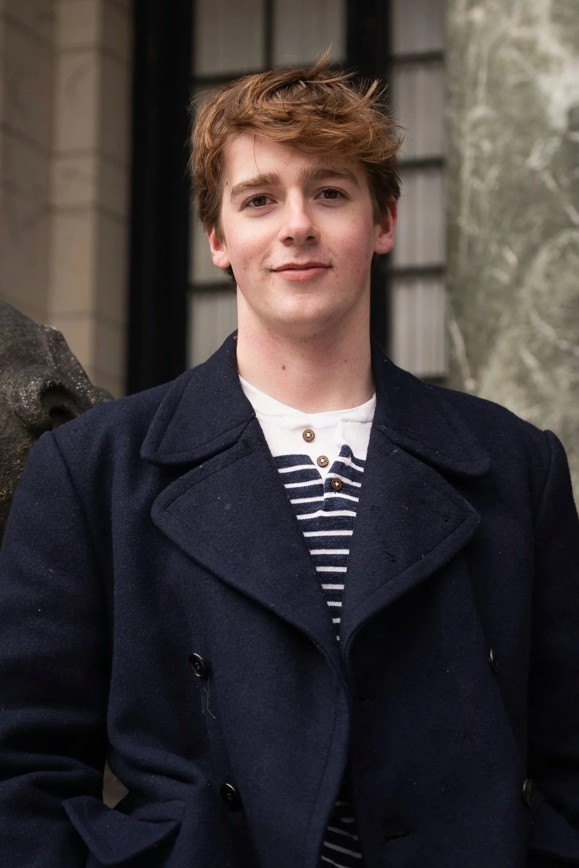 A young man with light skin, tousled light brown hair, and a slight smile, wearing a navy coat over a striped shirt, standing outdoors in front of a building with large windows and stone walls.