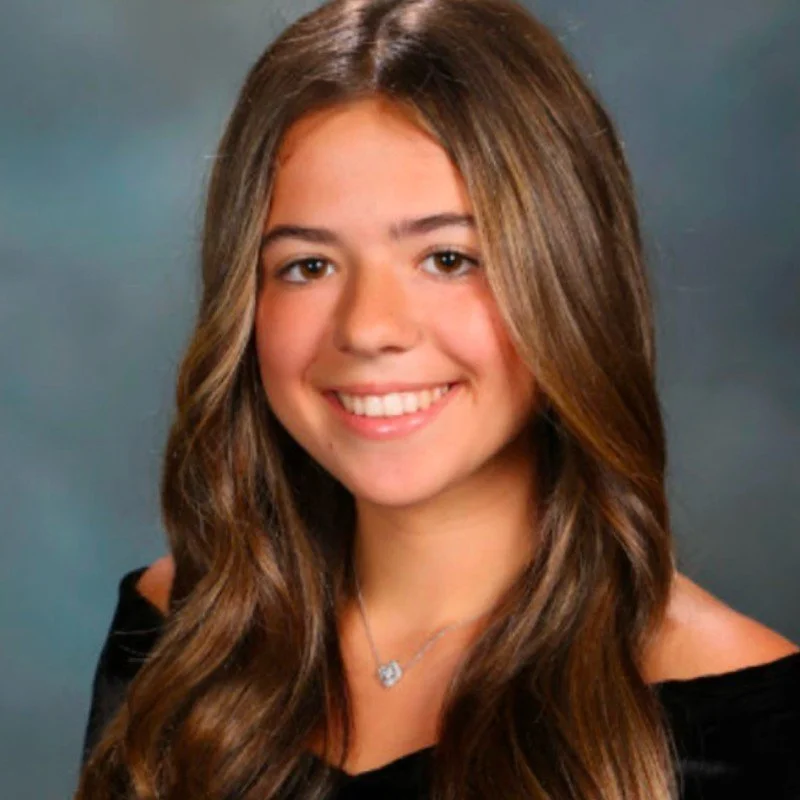 Portrait of a young woman with long, wavy brown hair, smiling, wearing a black top and a silver necklace, against a gray background.