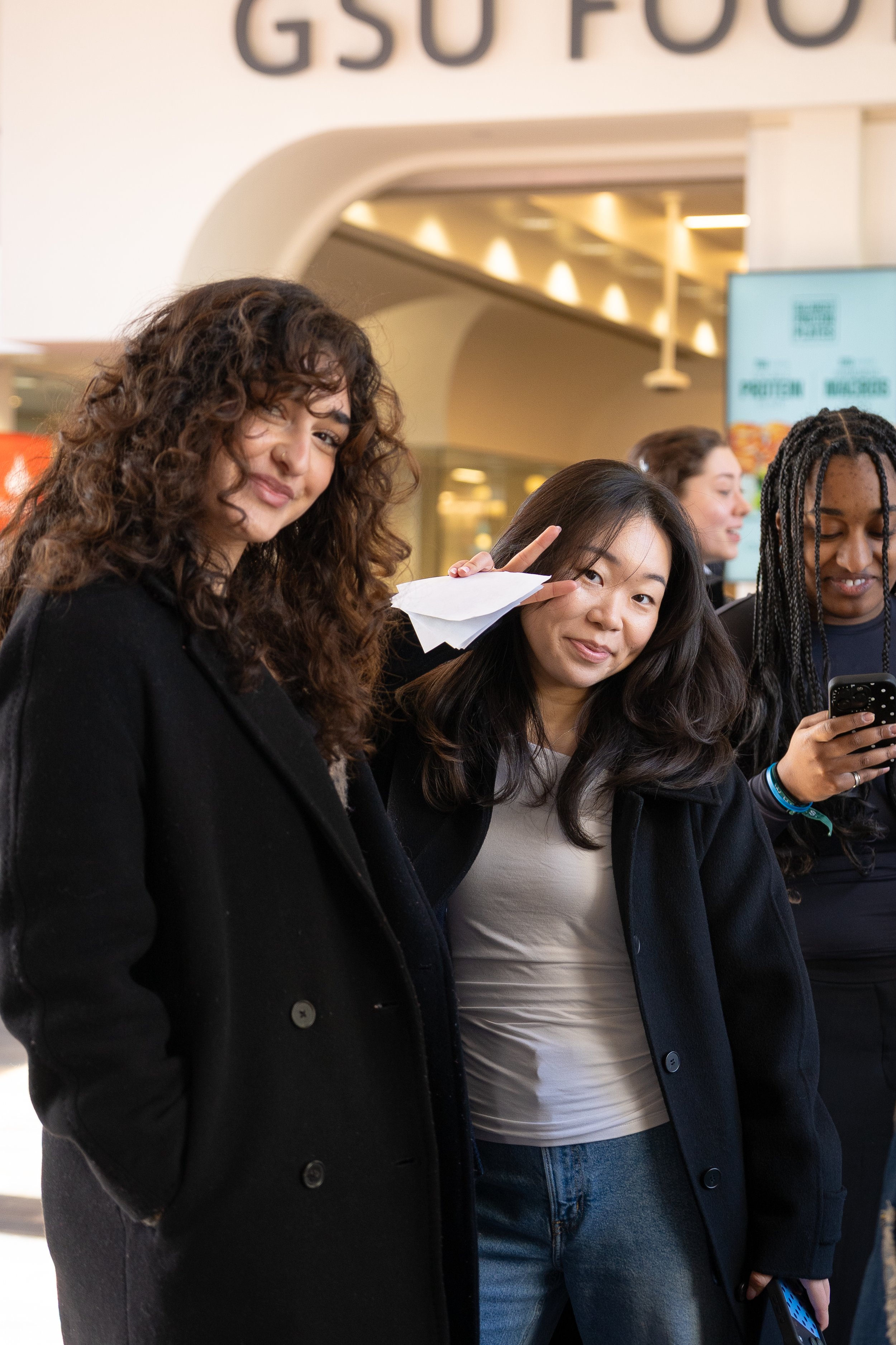 A group of women standing in line at a food court or cafeteria, with a sign reading 'GSO FOOD' in the background. One woman with curly hair is winking and smiling at the camera, while another woman with straight hair covers her eyes with her hand and smiles. Other women are visible in the background, some looking at their phones.