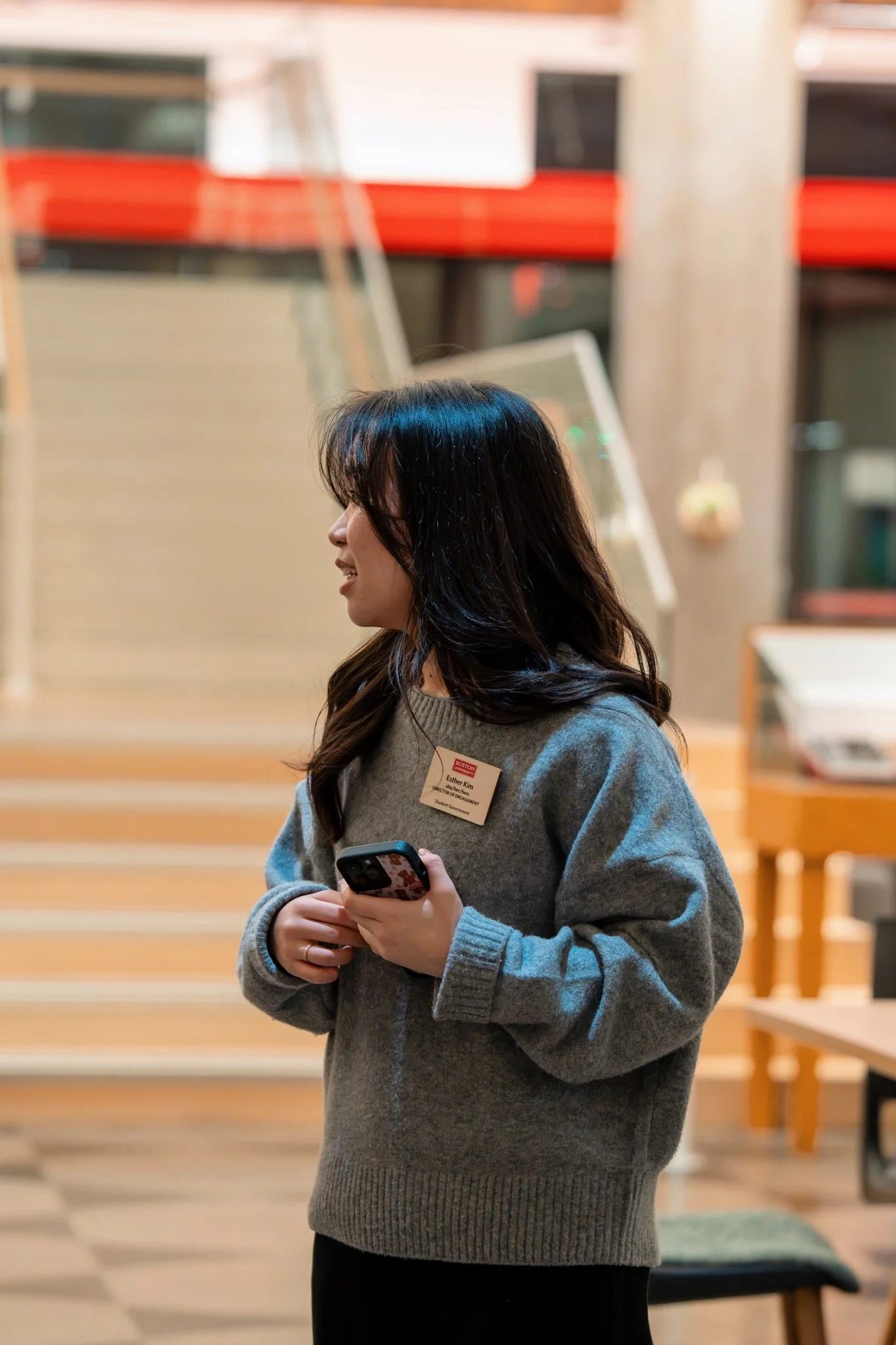A woman with long dark hair wearing a gray sweater holding a smartphone, standing in a room with wooden furniture and a colorful background.