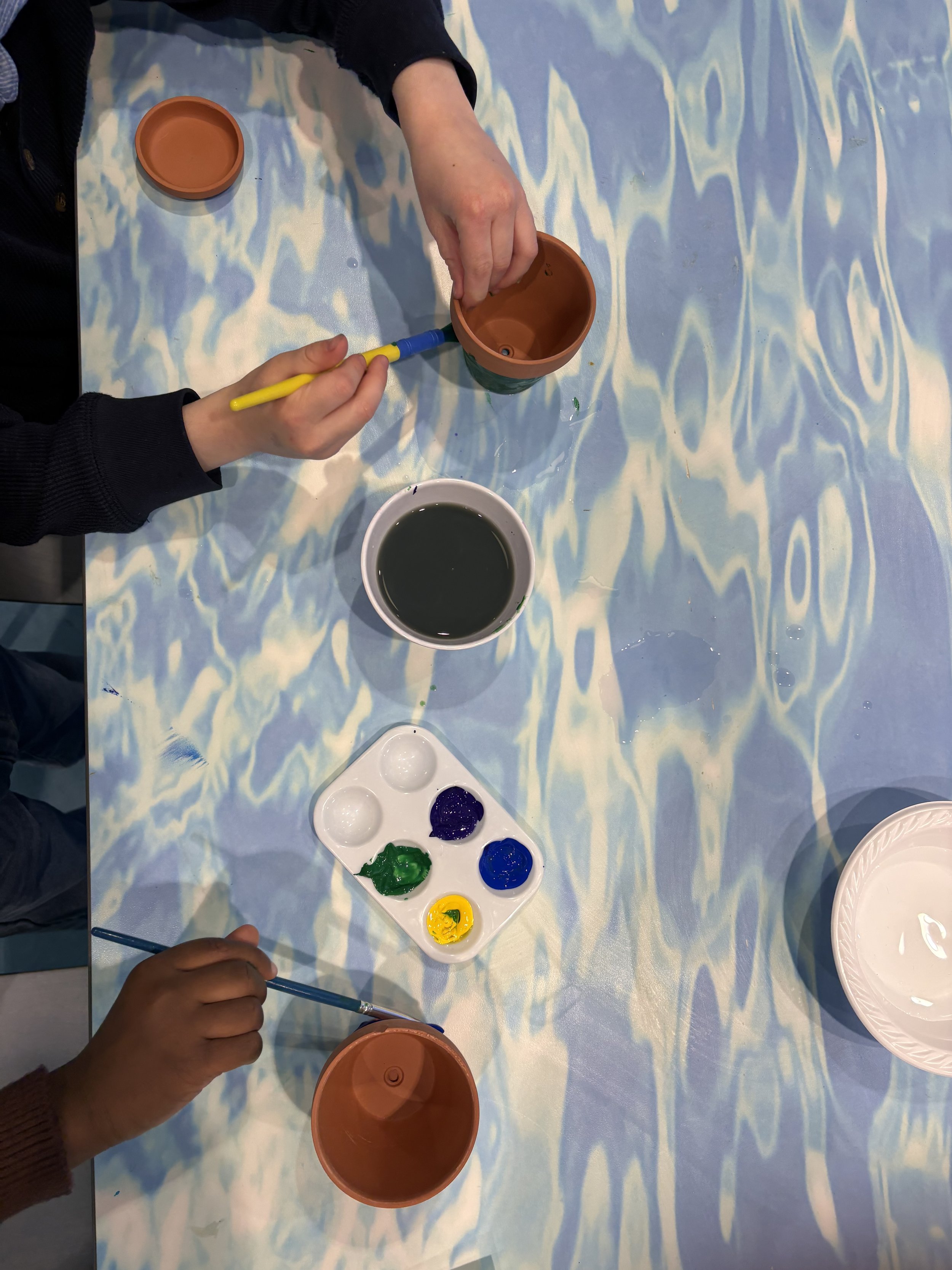Children are painting terra cotta pots with green, blue, and yellow paint on a blue tablecloth.