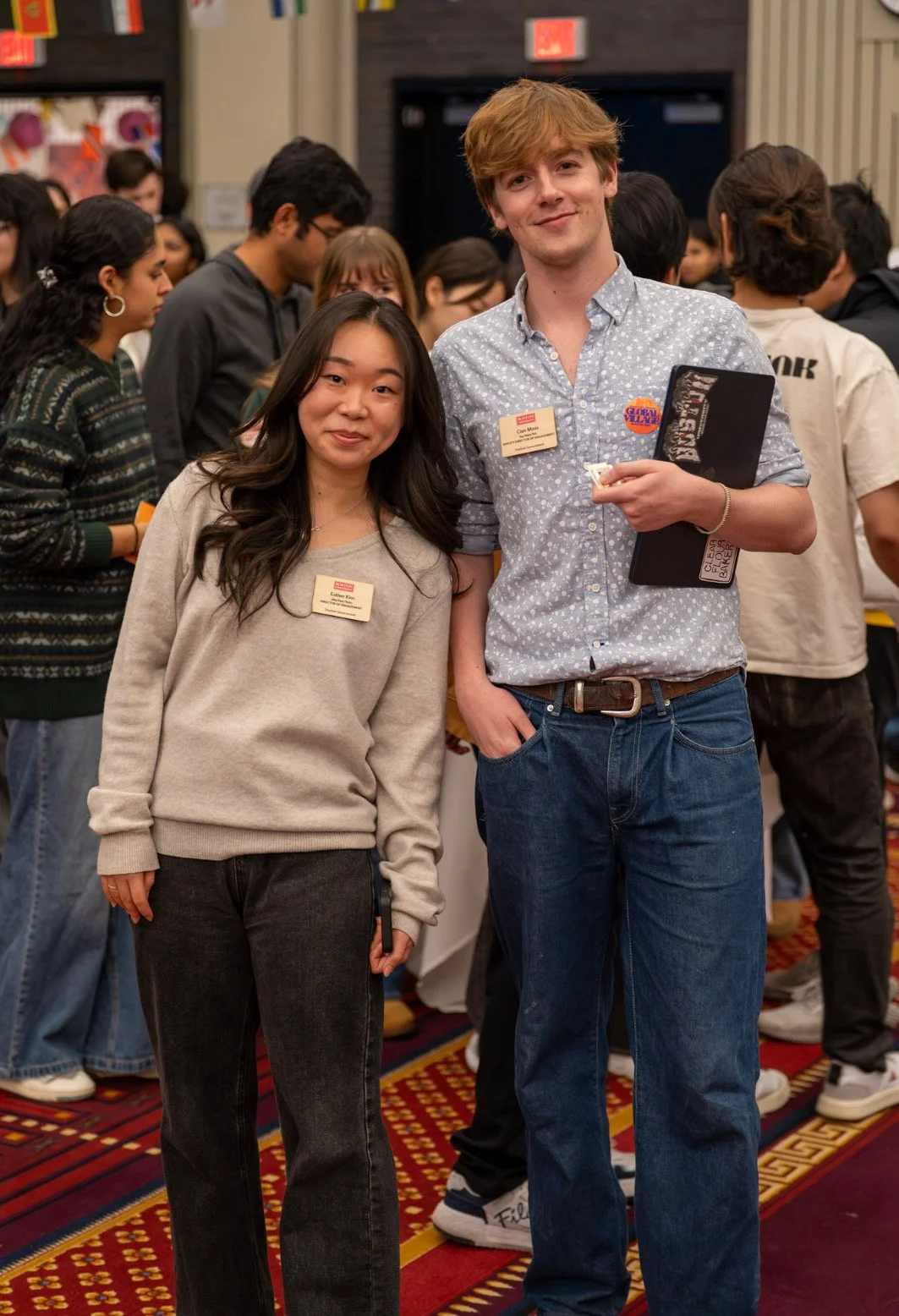 Two young individuals, a woman and a man, standing together at a busy indoor event with many other people in the background. The woman has long dark hair and is wearing a beige sweater and black jeans. The man has light brown hair, is wearing a light blue patterned shirt, and holding a folder and small items in his hand. Both are smiling and wearing name tags.
