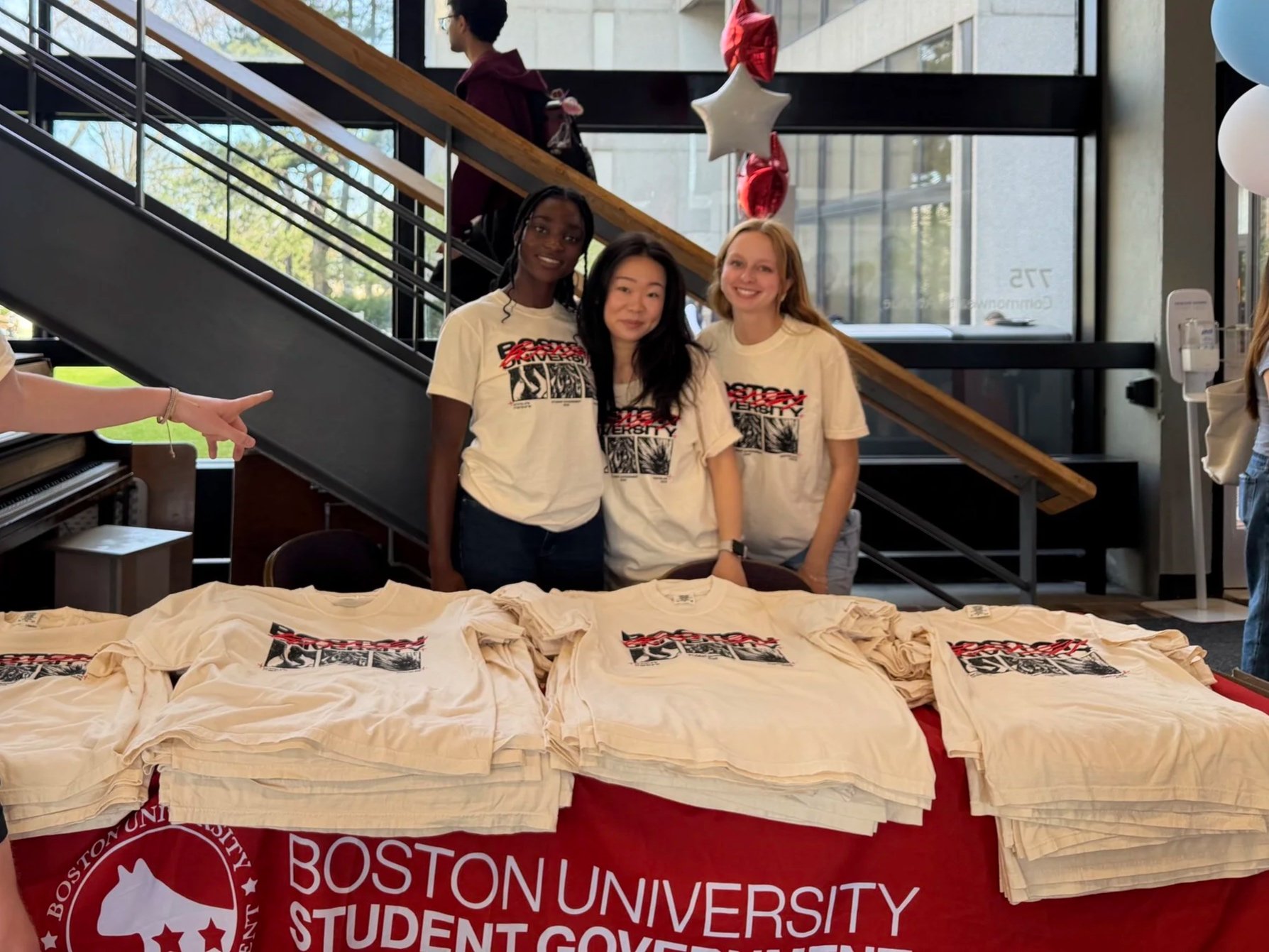 Three women standing behind a table covered with white T-shirts printed with a graphic design, inside a modern building with large glass windows. Balloons are hanging from the ceiling, and stairs are visible in the background. A person points towards the table.