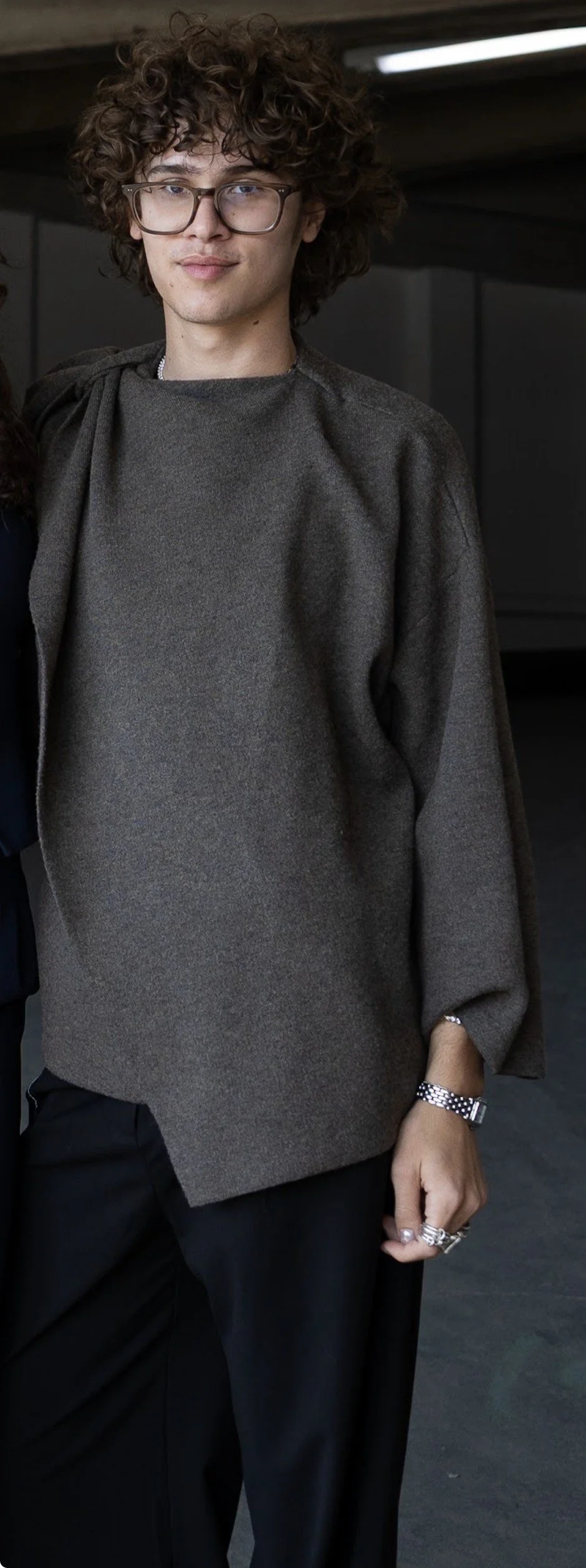 A person with curly hair and glasses wearing a dark gray top and black pants, standing indoors with a dark background.