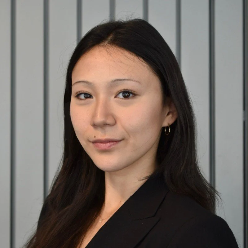 Portrait of a young woman with long dark hair, wearing a black blazer and small hoop earrings, standing against a modern gray-paneled background