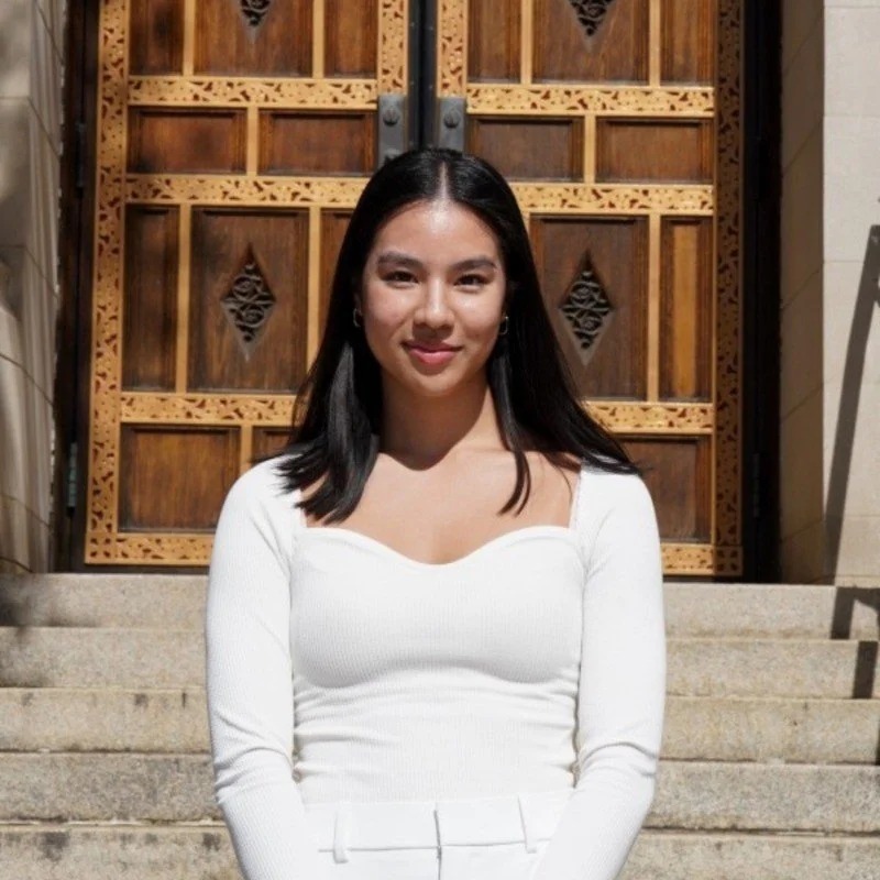 Young woman with black hair wearing a white long-sleeve top, standing on stone steps in front of wooden double doors.