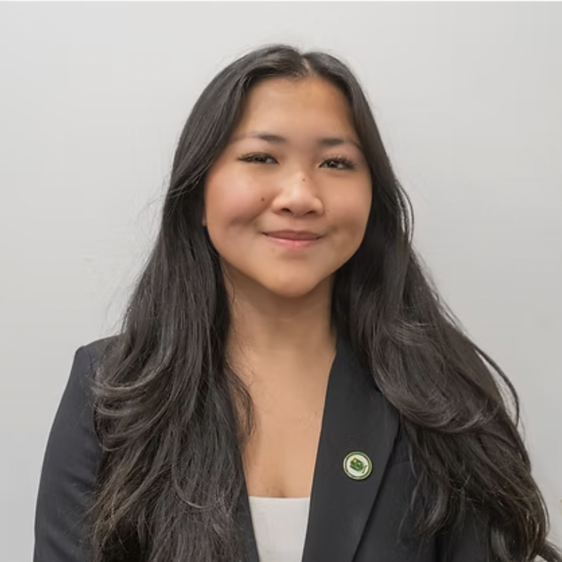 A woman with long black hair, smiling, wearing a black blazer with a green and white circular pin on it, and a white top underneath, standing against a plain light gray background.