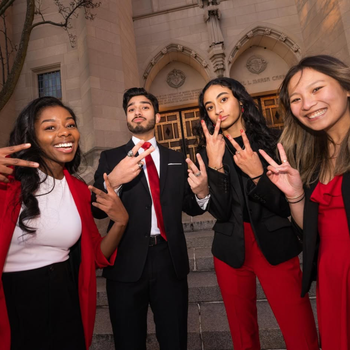 Four young adults posing in front of a building with an arched entrance, making peace signs and showing friendly expressions.